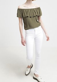 Oliven grøn off-the-shoulder top med rynket halsudskæring, parret med hvide slim-fit jeans og hvide sko med en sort sål.