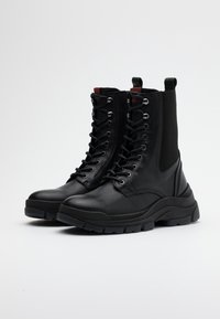 Bottes de combat en cuir noir avec une tige haute, un système de laçage et un panneau latéral texturé. Semelle épaisse et robuste pour une meilleure adhérence et durabilité.