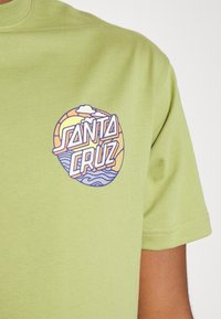 Světlé zelené bavlněné tričko s kruhovou grafikou obsahující nápis "SANTA CRUZ", vlny a mraky v pastelových barvách. Krátké rukávy a kulatý výstřih.