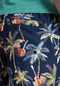 Plavky s tmavě námořnickým pozadím s tropickým vzorem palem a hudebních nástrojů. Obsahují zadní kapsu na knoflík.