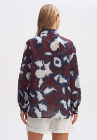Femme vue de dos portant une chemise ample à manches longues avec un imprimé floral bleu marine, blanc et bordeaux, associée à un short blanc retroussé.