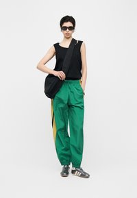 Άτομο με μαύρο αμάνικο τοπ, πράσινο και κίτρινο παντελόνι φόρμας, μαύρη τσάντα φάκελο, μαύρα αθλητικά παπούτσια και σκούρα γυαλιά ηλίου, στέκεται μπροστά σε λευκό φόντο.