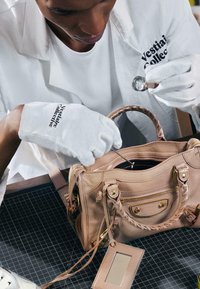 Personne portant des gants blancs et un uniforme Vestiaire Collective inspecte un sac à main en cuir beige à l'aide d'une loupe.