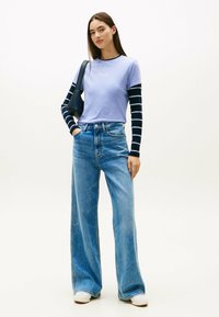 T-shirt viola chiaro con testo bianco, indossato sopra una maglietta a maniche lunghe a righe bianche e nere. Abbinato a jeans in denim a gamba larga a vita alta.