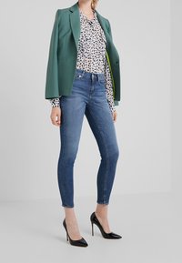 Mujer vistiendo jeans ajustados azules, zapatos de tacón negros, una blusa con un patrón blanco y negro, y una chaqueta verde sobre los hombros, de pie sobre un suelo gris.