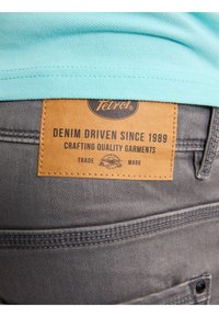 Tela de mezclilla gris con una etiqueta de cuero que dice "Tetrol DENIM DRIVEN SINCE 1989". El dobladillo superior es de algodón azul claro. Presenta costuras en el bolsillo trasero.