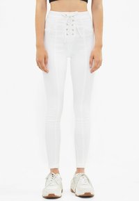 Vita högmidjade byxor med snörningsdetalj framtill och vertikala sömdetaljer, i kombination med ljusa sportskor.