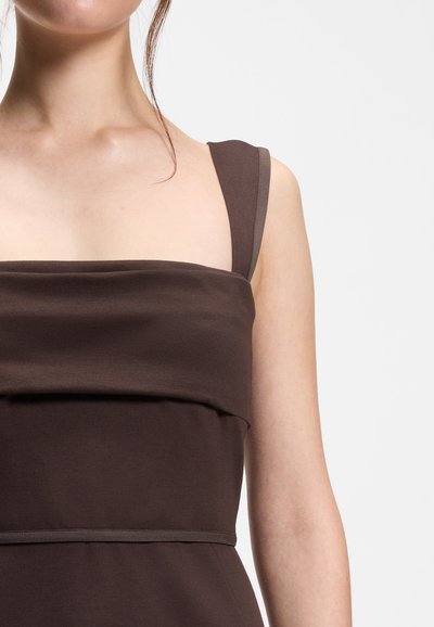 Jeune personne portant une robe marron foncé sans manches avec larges bretelles et un col plié, sur un fond uni.
