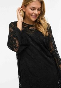 Femme souriante aux cheveux blonds portant une robe noire à manches longues en dentelle à fleurs, posant avec une main près de l'oreille.