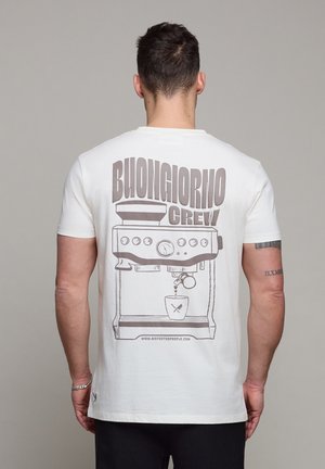 Man draagt een wit T-shirt met grote afbeelding van een koffiemachine en de tekst "Buongiorno Crew" op de achterkant, staand tegen een effen achtergrond.