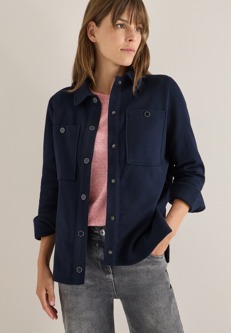 Chaqueta azul marino de botón con dos bolsillos frontales, tela texturizada y detalles en metal plateado. Llevada sobre un suéter rosa claro.