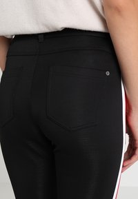 Schwarze dehnbare Jeans mit glatter Textur; verfügt über zwei Gesäßtaschen und seitliche Streifen in Rot und Weiß. Nähte sind entlang der Säume sichtbar.