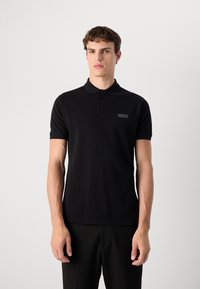 Polo noir en coton, avec un col boutonné, des manches courtes et un discret patch logo sur la poitrine gauche. Design épuré.