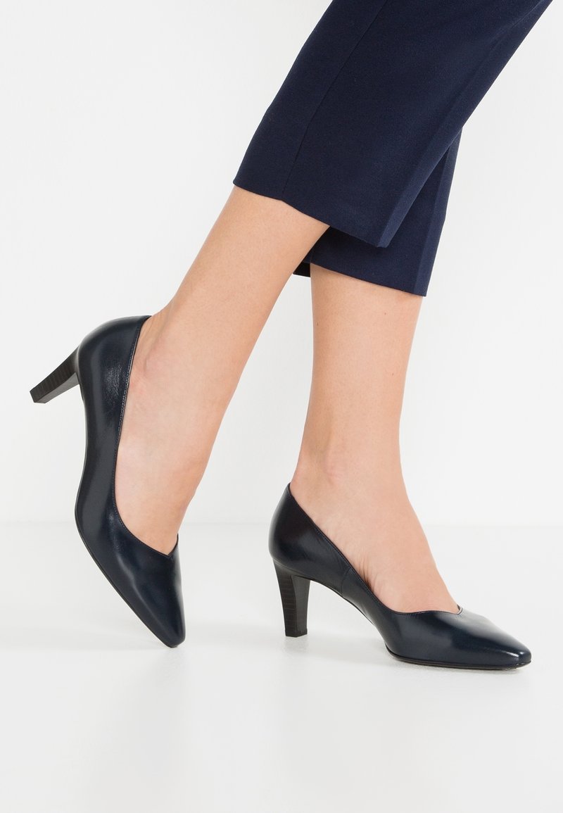 Femme portant des escarpins fermés bleu marine à talon moyen associés à un pantalon court bleu marine, debout sur un sol blanc devant un fond blanc.