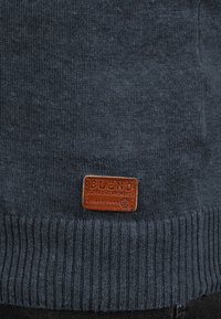 Dunkelblauer, gestrickter Pullover mit gerippten Bündchen und Saum, auf dessen Vorderseite ein braunes Lederetikett mit der Aufschrift "BLEND GLOBAL JEANSMAKER" angenäht ist.