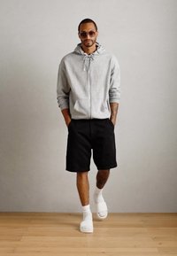 Šedá mikina na zips, čierne šortky, biele ponožky a šľapky. Hladký materiál, voľný strih a minimalistický dizajn, nosená s rukami v vreckách.