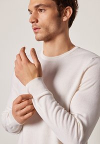 Weißer Strickpullover mit gerippten Bündchen und Ausschnitt, taillierte Passform, glatte Textur und subtiler Glanz, getragen von einem Modell, das Handdetails zeigt.