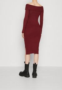 Robe bordeaux côtelée décolletée épaules avec manches longues, dotée de fentes latérales, associée à des bottes noires à talons épais.