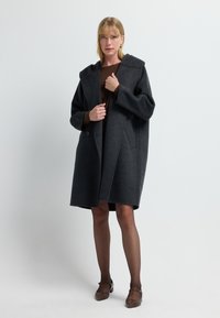 Abrigo de lana gris oscuro con capucha, diseño oversize, cierre frontal de botones y bolsillos laterales. Llevado sobre una camiseta marrón y combinado con zapatos marrones.