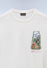 Camiseta blanca con un pequeño diseño impreso de una bolsa de red verde que contiene flores y frutas, con el texto "NAPAPIJRI" debajo.