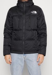 Sort polstret jakke med hætte, lynlåslukning foran, to brystlommer, The North Face-logo på venstre bryst og ribkanter ved ærmerne.