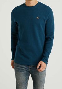 Långärmad t-shirt med rund hals i mörk teal, tillverkad av texturerad tyg, med en liten svart logotyp patch på vänster bröst.