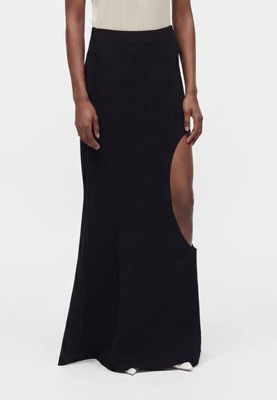 Black long skirt with a high side slit worn by a person with dark skin and a beige sleeveless top, white pointed shoes visible below.