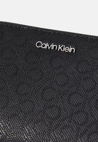 Melnā teksturētā maka ar ģeometrisko rakstu un sudraba "Calvin Klein" logotipa akcentu. Gluda virsma ar maigu spīdumu.