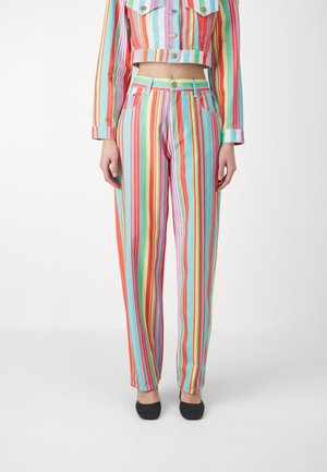 Femme portant un pantalon taille haute à rayures verticales multicolores avec une veste courte assortie et des chaussures noires, debout devant un fond blanc.