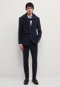 Marineblauer Anzug mit schmaler Passform, dazu passende Hosen, weißes Hemd und ein marineblauer Pullover, der um die Schultern gebunden ist. Schwarze Schuhe vervollständigen den Look.