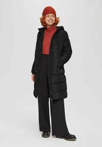 Schwarze Daunenjacke mit Kapuze und Reißverschluss, mit einem gesteppten Design. Kombiniert mit einem roten Rollkragenpullover und schwarzen Weit beinigen Hosen sowie einer gestrickten Mütze.