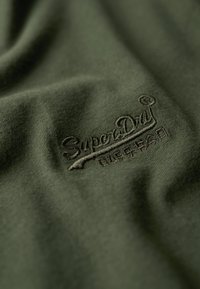 Tessuto di cotone verde scuro con un logo "SuperDry" in rilievo di tono leggermente più scuro, che mette in risalto una texture liscia e una leggera lucentezza.