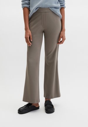 Person iført taupevidde bukser, lyseblå strikket trøje og sorte slip-on loafers, stående mod en ensfarvet baggrund.