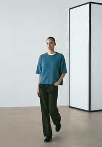 Vrouw die binnen loopt en een blauwe trui met korte mouwen, zwarte broek en zwarte schoenen draagt tegen een minimalistische wit-grijze achtergrond.