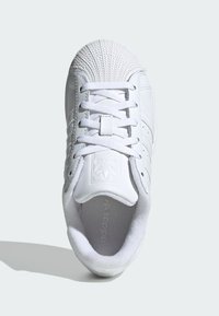 Bijela tenisica s teksturiranim vrhom izrađenim od školjke i glatkim kožnim tijelom, s ravnim vezicama, tri pruge kao ukrasom te podstavljenim unutrašnjim dijelom s logotipom.