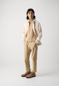 Beige wool sweater, cream button-up jacket, tan chinos, and brown shoes. Smooth textures, relaxed fit, subtle accents on the jacket.