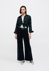 Πουκάμισο σε πράσινο και ναυτικό καρό με κουμπιά, δεμένο στη μέση, συνδυασμένο με ψηλόμεσα μαύρα παντελόνια από κορδόνι με φαρδιά πόδια και μαύρα αθλητικά παπούτσια.