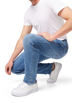 Homme agenouillé sur un genou, portant des baskets blanches, un jean bleu et un t-shirt blanc uni sur fond blanc.