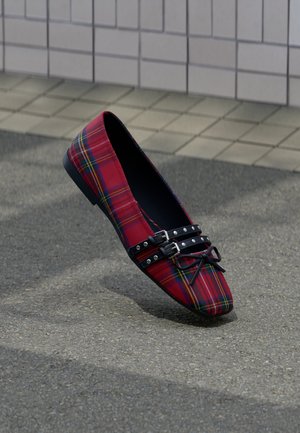 Czerwony płaski but w kratę tartan z zaostrzonym noskiem, czarne podwójne paski z ćwiekami i kokardą, ustawiony na asfalcie blisko ściany z płytek.