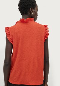 Rotes, strukturiertes ärmelloses Oberteil mit rüschierten Schulterakzenten und einem hohen Ausschnitt, entworfen für eine figurbetonte Silhouette.