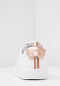 Białe sneakersy z wytłoczonym logo, różowozłota pięta z logo krokodyla oraz różowozłota i biała paskowana pętla.