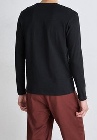 Black long-sleeve shirt made of cotton, with a round neck and slim fit, paired with brown trousers, viewed from the back.