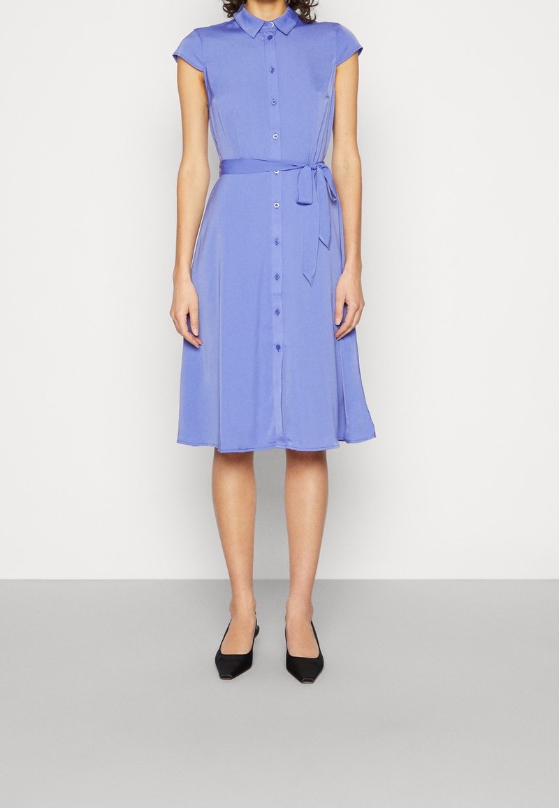 Robe boutonnière mauve clair avec des manches courtes à volants, une taille ceinturée et une jupe évasée. Des chaussures noires à bout pointu complètent le look.