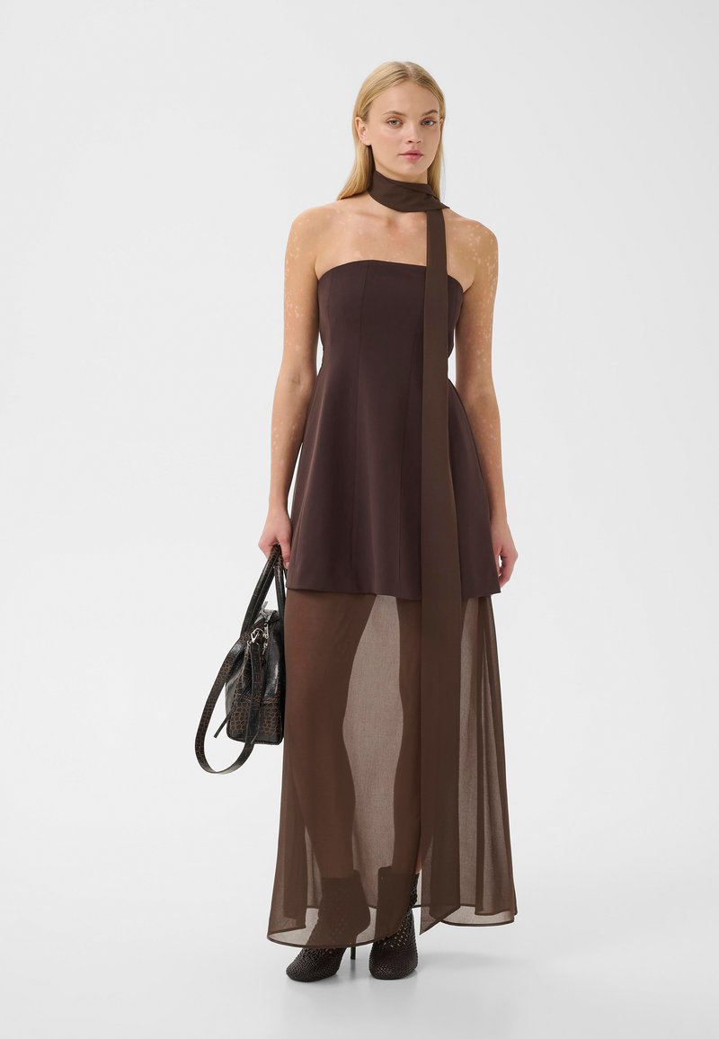 Femme blonde portant une robe bustier marron foncé avec une longue jupe transparente, foulard assorti autour du cou, tenant un sac à main noir texturé, debout sur fond blanc.