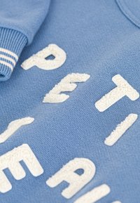 Modrá mikina s texturovaným krémovým nápisem. Hladké manžety s bílými pruhy, měkký materiál s hladkým povrchem.