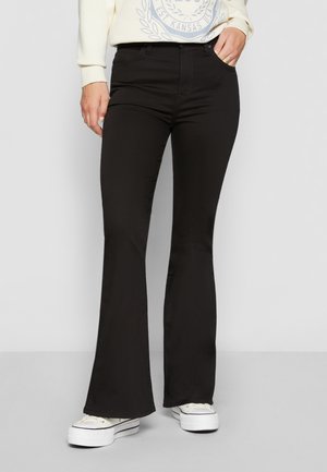 Personne portant un pantalon noir évasé, des baskets blanches à plateforme et une chemise à manches longues de couleur claire avec un motif imprimé.