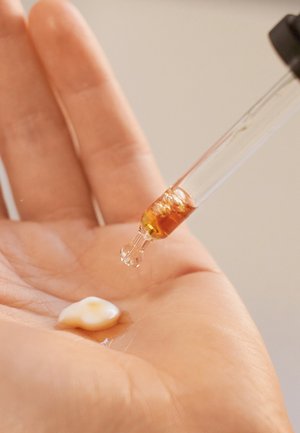 Hand mit Lotion und bernsteinfarbener Flüssigkeit, die aus einer Pipette auf die Handfläche gegeben wird.