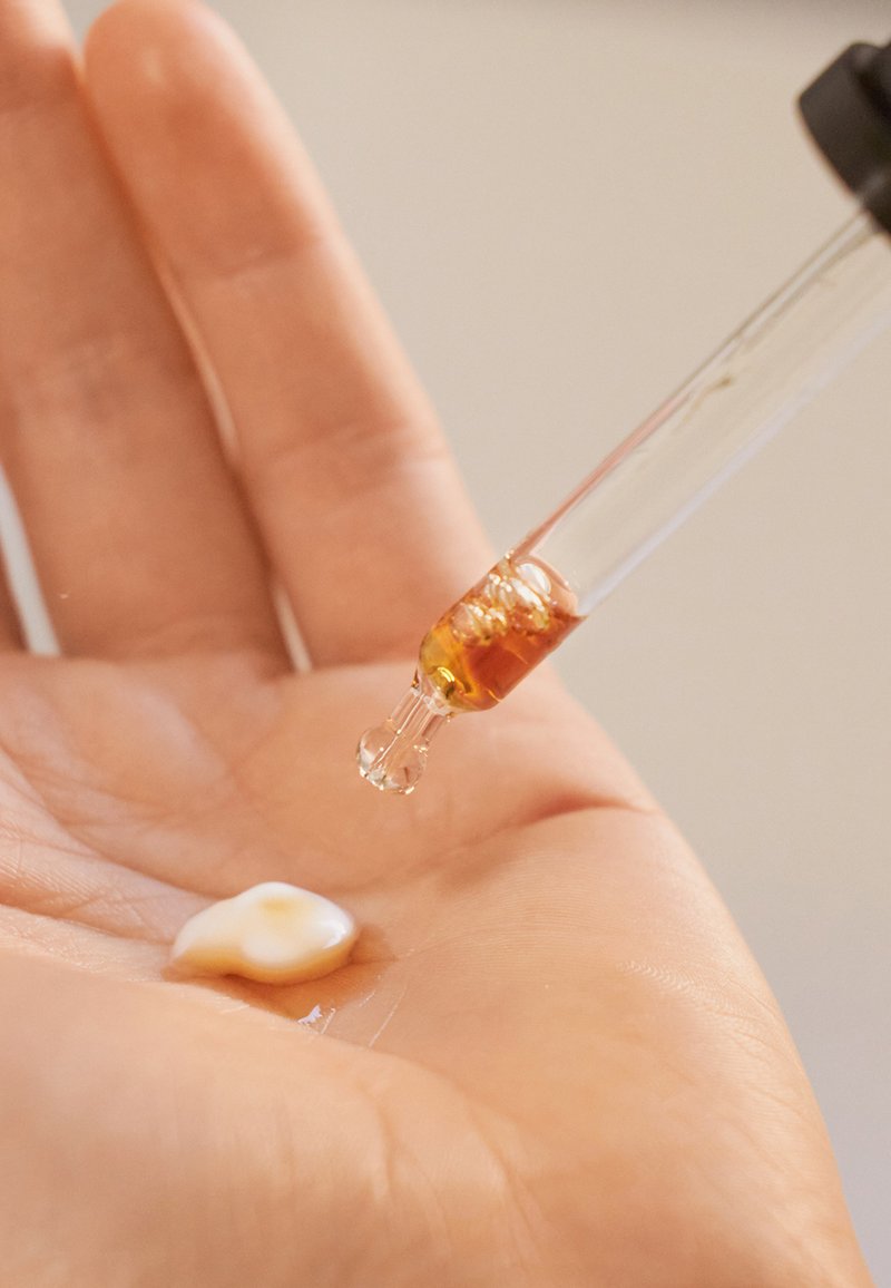 Hand mit Lotion und bernsteinfarbener Flüssigkeit, die aus einer Pipette auf die Handfläche gegeben wird.