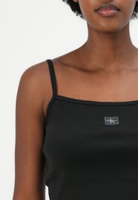 Black ribbed tank top with thin straps featuring a small gray Calvin Klein logo label at the center front. Smooth texture.