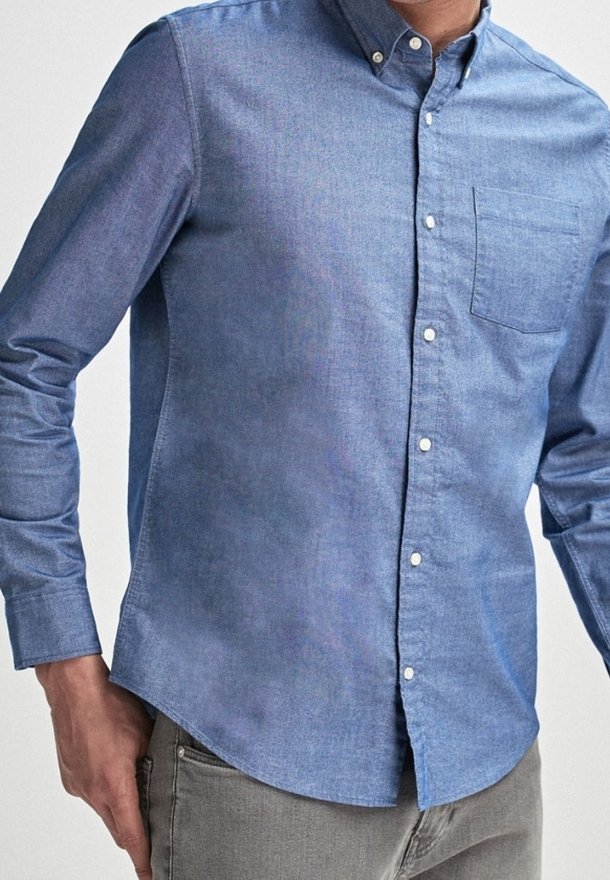 Man in een lichtblauwe overhemd met knopen en een borstzak, met grijze broek, staande met één hand dicht bij zijn heup.